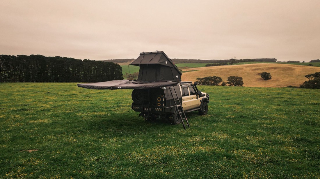 A LandCruiser in a green paddock with a black OZTREK4x4 rooftop tent open on the roof and the awning out